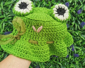Könnte beinhalten: Grüne gehäkelte Frosch-Mütze mit weißen und schwarzen Augen und einem grünen Band.