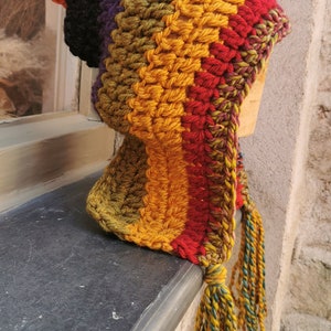 K&ouml;nnte beinhalten: Eine bunte H&auml;kelm&uuml;tze mit einem langen Quasten. Die M&uuml;tze ist aus Streifen von orangefarbenem, schwarzem, violettem, gr&uuml;nem, gelbem und rotem Garn gefertigt.