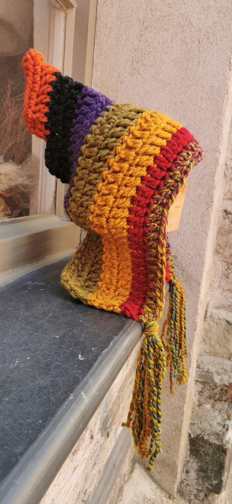 K&ouml;nnte beinhalten: Eine bunte H&auml;kelm&uuml;tze mit einem langen Quasten. Die M&uuml;tze ist aus Streifen von orangefarbenem, schwarzem, violettem, gr&uuml;nem, gelbem und rotem Garn gefertigt.