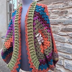 May include: A colourful, handmade crochet cardigan in a granny square design. The open-front garment features orange, green, purple, blue, and brown squares. The cardigan is displayed on a mannequin against a stone wall background.