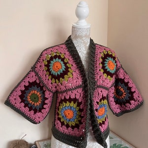 May include: A pink crocheted cardigan with brown trim and colourful floral motifs. The cardigan has a shawl collar and three-quarter sleeves.