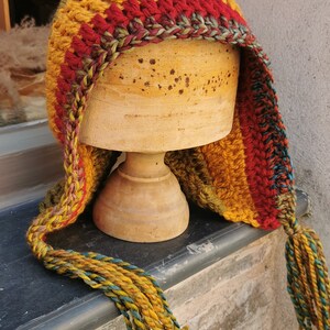 K&ouml;nnte beinhalten: Eine bunte H&auml;kelm&uuml;tze mit einem langen Quasten. Die M&uuml;tze ist aus Streifen von rotem, orangefarbenem, gelbem, gr&uuml;nem und violettem Garn gefertigt. Die M&uuml;tze wird auf einem Holzst&auml;nder pr&auml;sentiert.