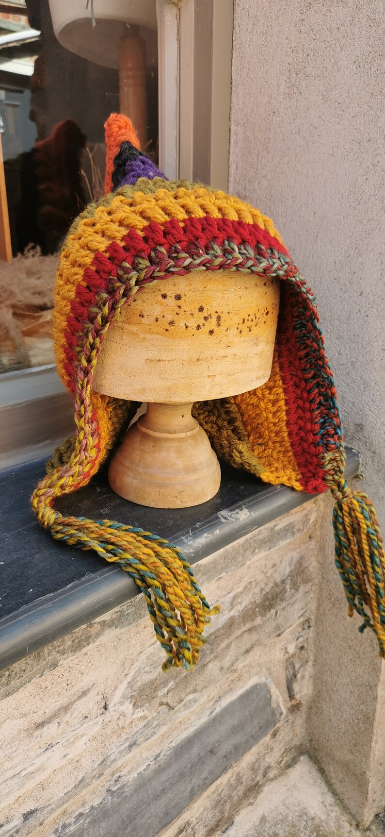 K&ouml;nnte beinhalten: Eine bunte H&auml;kelm&uuml;tze mit einem langen Quasten. Die M&uuml;tze ist aus Streifen von rotem, orangefarbenem, gelbem, gr&uuml;nem und violettem Garn gefertigt. Die M&uuml;tze wird auf einem Holzst&auml;nder pr&auml;sentiert.