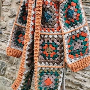 May include: A crocheted cardigan featuring a granny square pattern in shades of orange, green, brown, and grey. The cardigan is open at the front and has long sleeves.