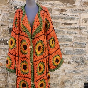 May include: A long, orange and green crocheted cardigan with a sunflower pattern. The cardigan is on a mannequin wearing a green beanie. The sign behind the mannequin reads "Ty Custom House Shop Siopa Gallery Oriel".