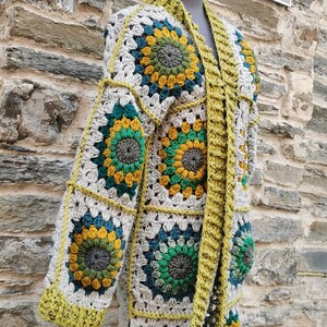 May include: A long, crocheted cardigan with a green, yellow and blue floral pattern. The cardigan is open at the front and has a long, flowing silhouette. It is displayed on a mannequin wearing a matching green hat.