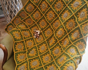 Könnte beinhalten: Eine gehäkelte Wolldecke mit einem Sonnenblumenmuster in Gelb- und Grüntönen. Die Decke ist aus einem strukturierten Garn gefertigt und hat eine quadratische Form.