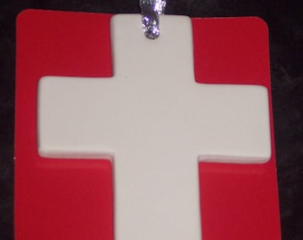 May include: A white ceramic cross ornament with rounded edges, suspended by a shimmering silver ribbon. The cross is positioned against a solid red rectangular backing card, creating a high-contrast display of white, red, and silver elements.