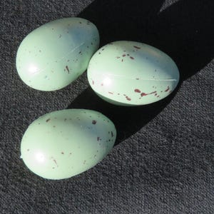 May include: Three light green, speckled eggs are arranged on a dark gray surface. The eggs have a smooth, glossy finish and are dotted with small, reddish-brown markings. The eggs are oval-shaped and appear to be made of a hard material.