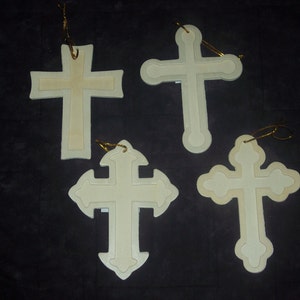 May include: Four off-white cross ornaments with gold string hangers. Each cross has a layered design with a lighter center. The crosses vary in shape, with some having rounded edges and others more angular.