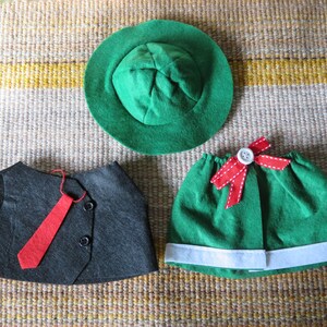 May include: A green felt hat, a dark gray vest with a red tie, and a green skirt with a white trim and a red bow. The items are arranged on a textured, neutral-toned surface.