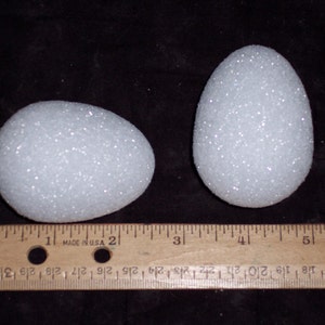 May include: Two white, glitter-covered decorative eggs of different sizes are displayed on a black surface. A wooden ruler with inch markings is placed next to the eggs for scale. The eggs have a textured, sparkling appearance.
