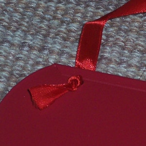 May include: A red, heart-shaped item with a red satin ribbon attached. The ribbon is knotted through a small hole in the item. The background is a textured, neutral-colored surface.