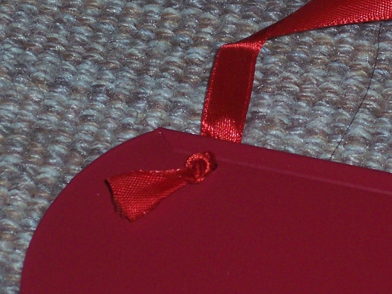 May include: A red, heart-shaped item with a red satin ribbon attached. The ribbon is knotted through a small hole in the item. The background is a textured, neutral-colored surface.