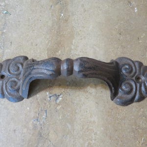 May include: A dark brown, cast iron drawer pull featuring an ornate, scrolled design on both ends. The handle has a central raised section and two circular mounting holes, one on each side, for attachment to a surface or piece of furniture.