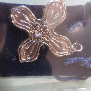 May include: A silver-colored, four-petal flower-shaped pendant with a central floral design. The pendant is attached to a small, dark blue velvet pouch. The text "Petit sac souvenir en velvet compris" is visible.