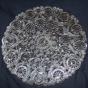 May include: A silver-colored paper doily with a circular design featuring intricate, repeating patterns of small circles and swirls.