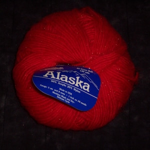 May include: A vibrant red ball of Alaska yarn with a blue label. The label indicates the yarn is 60% acrylic and 20% nylon, made in the USA, and contains 130 yards. The yarn is suitable for machine washing and drying on a delicate cycle.
