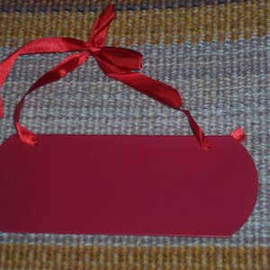 May include: A solid red, oval-shaped tag with rounded edges and a red satin ribbon tied in a bow at the top. The tag is blank, ready for personalization. The background is a textured, multi-colored fabric.