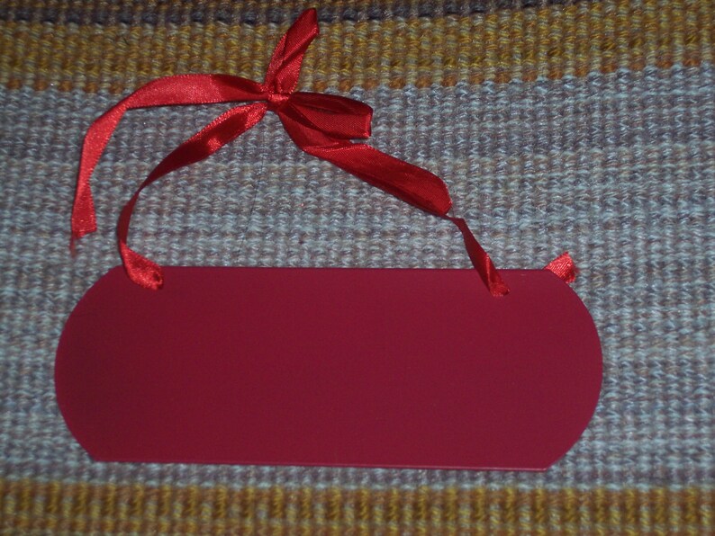 May include: A solid red, oval-shaped tag with rounded edges and a red satin ribbon tied in a bow at the top. The tag is blank, ready for personalization. The background is a textured, multi-colored fabric.