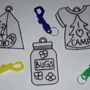 May include: A collection of camping-themed keychains. Designs include a tent with the word "zzz", a jar labeled "BUGS", and a shirt with "I LOVE CAMP". Keychains are attached to blue, yellow, and green clips.