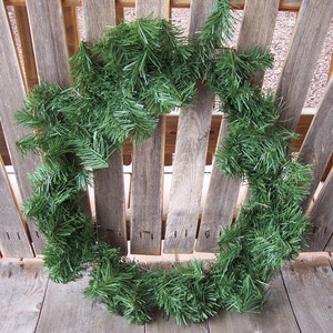 May include: A circular, evergreen wreath composed of dense, green artificial pine needles. The wreath is displayed against a weathered, wooden background, suggesting a rustic or natural aesthetic. Ideal for seasonal home decor.