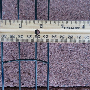 Puede incluir: Una estructura de marco de alambre verde oscuro colocada debajo de una regla de madera Fiskars. La regla muestra que el espacio entre los alambres mide aproximadamente 1,9 cm entre las barras verticales, extendi&eacute;ndose por la regla.