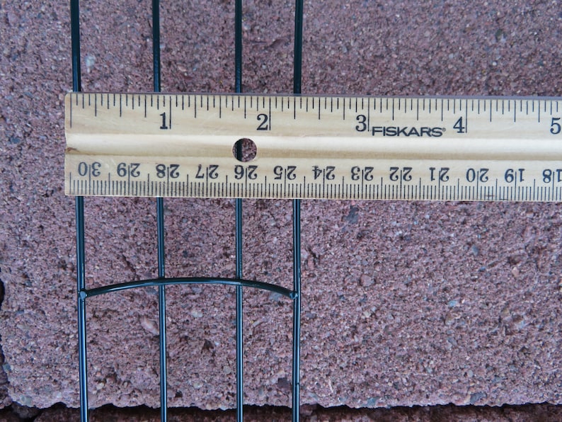 Puede incluir: Una estructura de marco de alambre verde oscuro colocada debajo de una regla de madera Fiskars. La regla muestra que el espacio entre los alambres mide aproximadamente 1,9 cm entre las barras verticales, extendi&eacute;ndose por la regla.