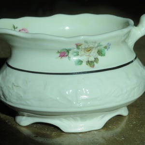May include: Cream-colored ceramic creamer with a floral design. The creamer has a scalloped rim, a handle, and a black band around the middle. The floral design includes white, pink, and blue flowers. The base has a decorative, leaf-like design.