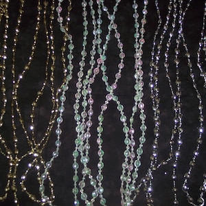 May include: Several strands of beaded garlands in gold, iridescent green and pink, and silver hang against a dark background. The beads are small and round, reflecting light.