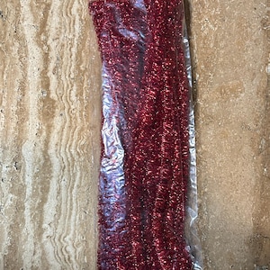 May include: A package of red tinsel garland. The tinsel is a vibrant, shiny red color and appears to be tightly packed within a clear plastic bag. The garland is likely used for holiday or craft decorations.