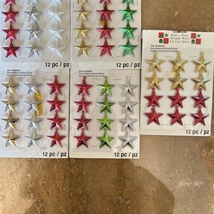 Puede incluir: Varios paquetes de adornos navideños en forma de estrella en rojo, plateado, dorado y verde. Cada paquete contiene 12 adornos. El embalaje incluye el texto "Merry Minis" y "Star Ornaments".