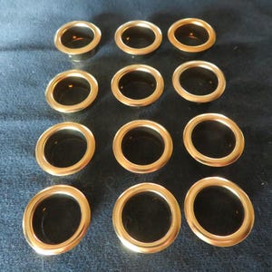 May include: A collection of twelve gold-colored metal grommets, arranged in three rows on a dark surface. Each grommet has a circular shape with a smooth, shiny finish. The inner circle is dark, creating a contrast with the gold exterior.