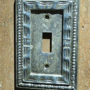 May include: A rectangular, antique-style light switch cover with a silver and blue finish. The cover features a decorative beaded border and a central opening for the switch. The surface has a textured, aged appearance.