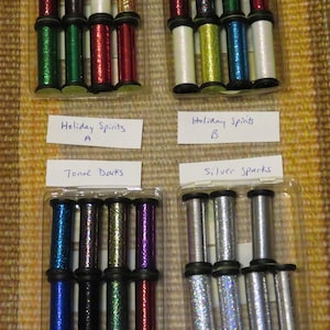 May include: A collection of spools of glitter thread in various colors, arranged in clear plastic containers. The colors include red, green, silver, gold, blue, and purple. The image includes labels that read "Holiday Spirits A", "Holiday Spirits B", "Tonal Darks", and "Silver Sparks".
