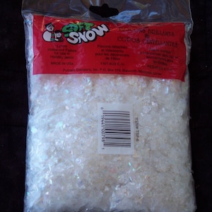 May include: A clear plastic bag filled with iridescent white flakes, labeled "Soft Snow." The red label features a snowman graphic and text in English and Spanish, indicating the product is for holiday decor. Made in the USA.