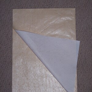 May include: A rectangular sheet of light tan, textured paper with a smooth, white, semi-transparent layer partially peeled back from the top left corner, revealing the contrasting textures and colors of the two distinct paper surfaces.