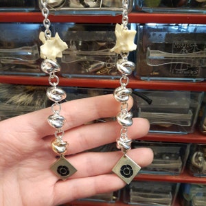 May include: Two silver chains with a bone charm and a silver flower charm at the end. The chains have silver knots and are hanging from a hand.