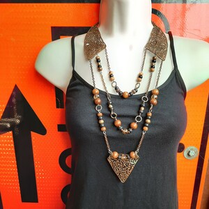 Peut inclure: Un collier à plusieurs rangs avec des perles brunes et noires et un pendentif triangulaire au design complexe. Le collier est en métal et en bois.