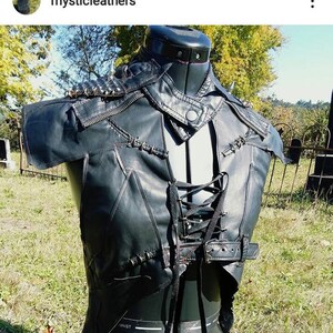 May include: Black leather vest with a lace-up front and studded shoulder accents. The vest features a zip closure, a buckled belt, and decorative stitching. The vest is displayed on a black mannequin in an outdoor setting.
