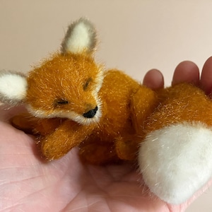 Könnte beinhalten: Eine kleine, orangefarbene und weiße Fuchsstatuette liegt in einer Hand. Der Fuchs hat geschlossene Augen, eine schwarze Nase und weiß bespitzte Ohren. Der flauschige Schwanz des Fuchses ist weiß. Die Figur besteht aus weichem Plüschmaterial.