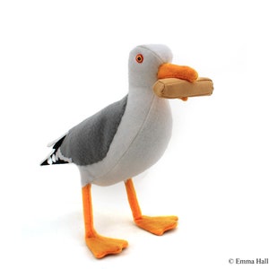 May include: A plush gray and white seagull toy holding a brown rectangle in its beak. The seagull has orange feet and a yellow beak.