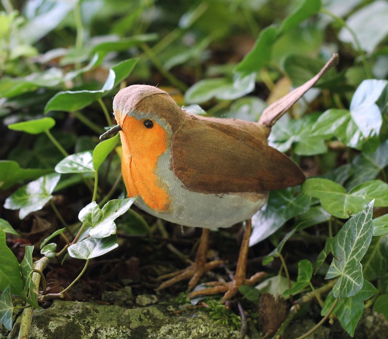 May include: A brown and gray felt robin with an orange breast, perched on a bed of green leaves.