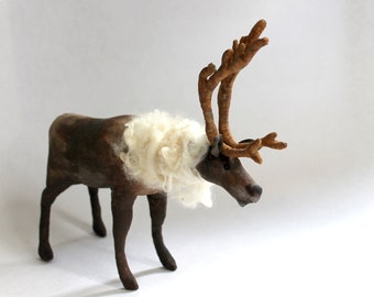May include: A handcrafted reindeer figurine with a brown body, dark brown legs, and large, textured antlers. The reindeer has a fluffy, cream-colored wool mane. The figurine is standing on a white surface.
