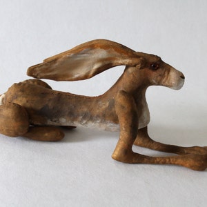 May include: A brown and white hare sculpture with long ears and a long, slender body. The hare is lying on its side with its head turned to the right. The sculpture is made of fabric and has a realistic appearance.