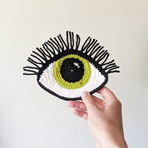 Könnte beinhalten: Ein gehäkeltes Auge mit schwarzen Wimpern, einem weißen Außenring, einem grünen Mittelring und einer schwarzen Mitte.
