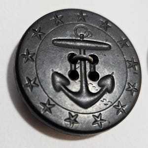 May include: A dark gray button with an anchor design. The button features an anchor in the center, surrounded by a ring of stars. The button has two holes for sewing and is likely made of metal.