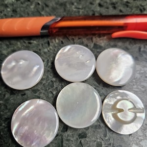 May include: Six round, iridescent mother-of-pearl buttons. The buttons are white with subtle pink and blue hues. One button has a two-hole shank. An orange pen is in the background.