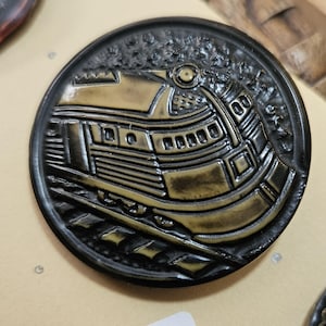 May include: A round, black metal medallion with a raised relief of a vintage train. The train is detailed in a gold color, with visible windows, a smokestack, and a prominent front. The medallion has a textured border.