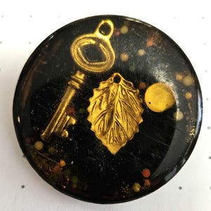 May include: A round, black button with gold accents. The button features a key, a leaf, and a circle. The button is covered in small, colorful glitter.